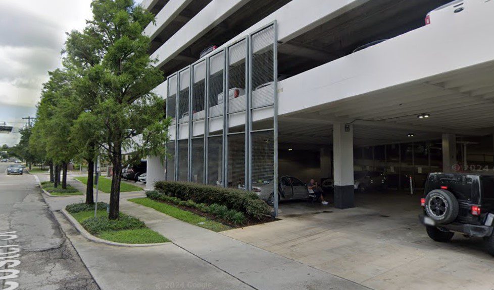 A multi-level parking garage with open sides, adjacent to a sidewalk with green grass, bushes, and trees. Several cars are parked inside, and a person stands near a silver vehicle.