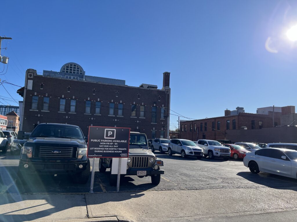 A parking lot filled with various cars on a sunny day, with a sign indicating public parking availability in front. A large brick building with tall windows is in the background against a clear blue sky.