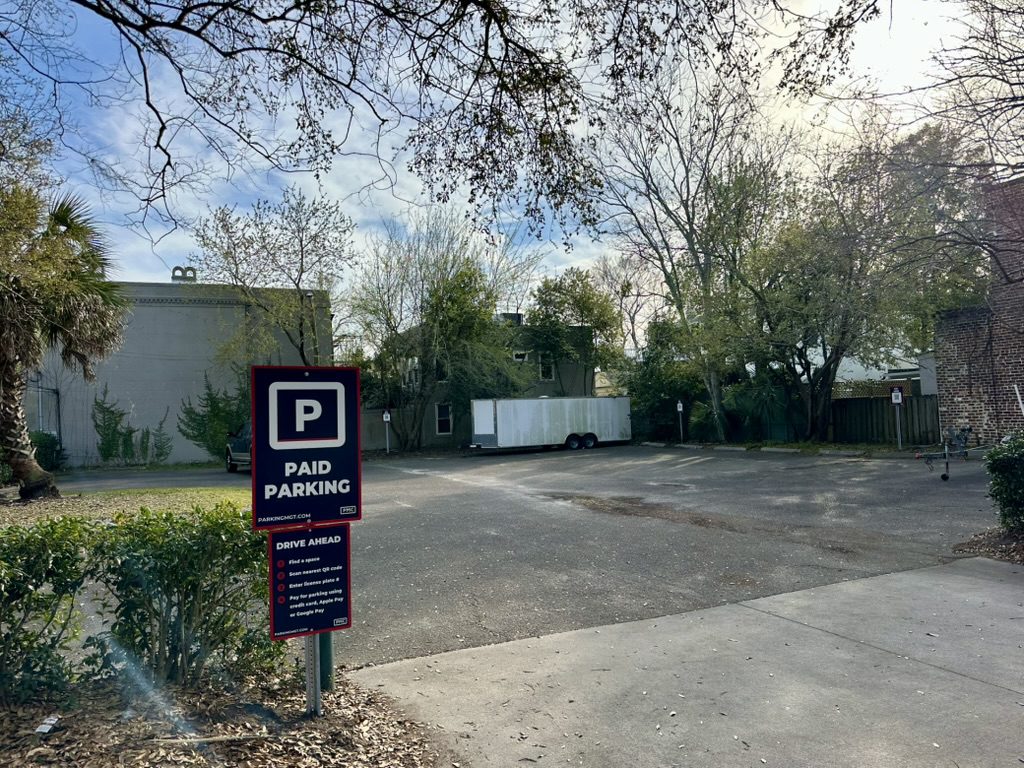 Outdoor Parking Management Company parking lot with a "Paid Parking" sign with the PMC logo in the foreground, surrounded by trees, shrubs, and a few buildings.