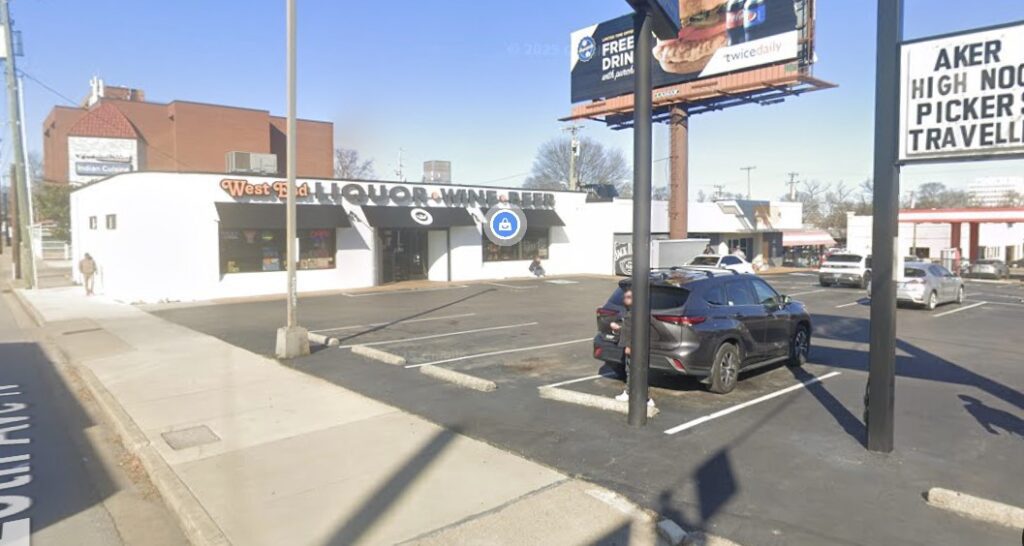 A liquor, wine, and beer store with a white exterior and large sign sits on a street corner. A few cars are parked in front, and a large billboard stands nearby advertising a free drink offer.