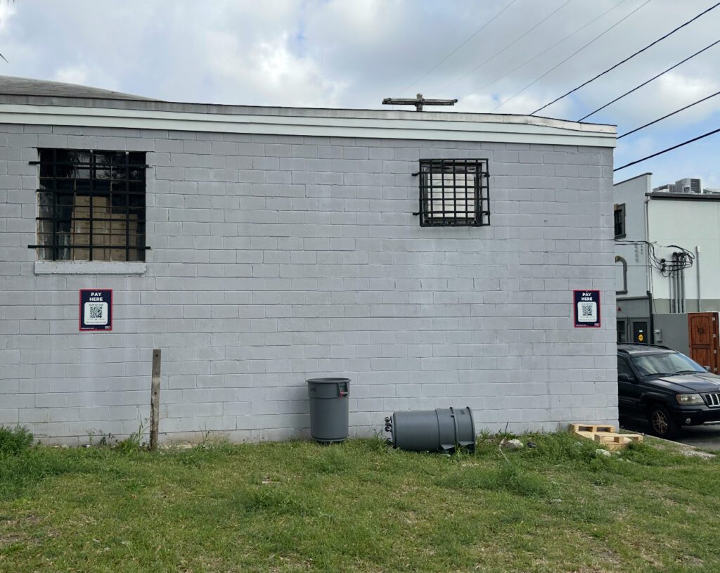A gray cinder block building with two barred windows. Two PMC signs with QR codes are posted on the wall. A car and utility poles are visible in the background.
