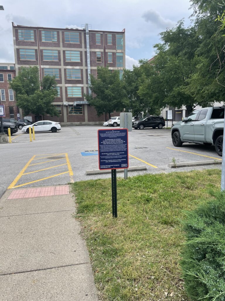 A small parking lot with a few cars, bordered by a sidewalk and grass. A prominent sign with text is posted near the entrance. In the background, there is a multi-story brick building and several trees.