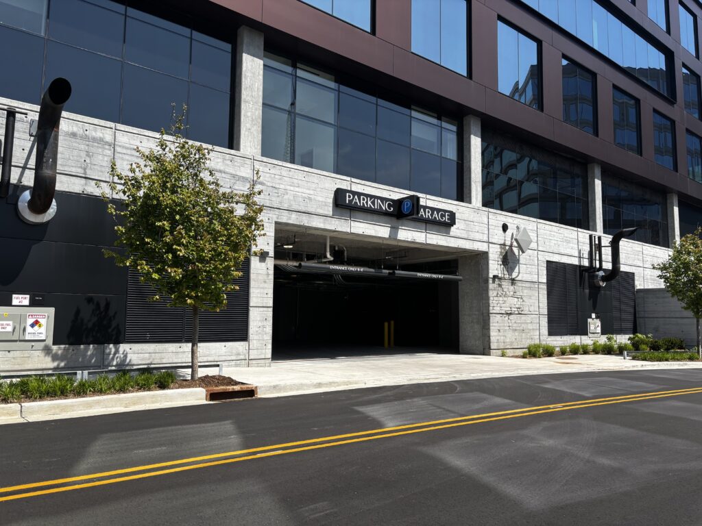 Entrance to a modern parking garage in a commercial building, with a "Parking Garage" sign above, trees on either side, and a road in the foreground under a sunny sky.