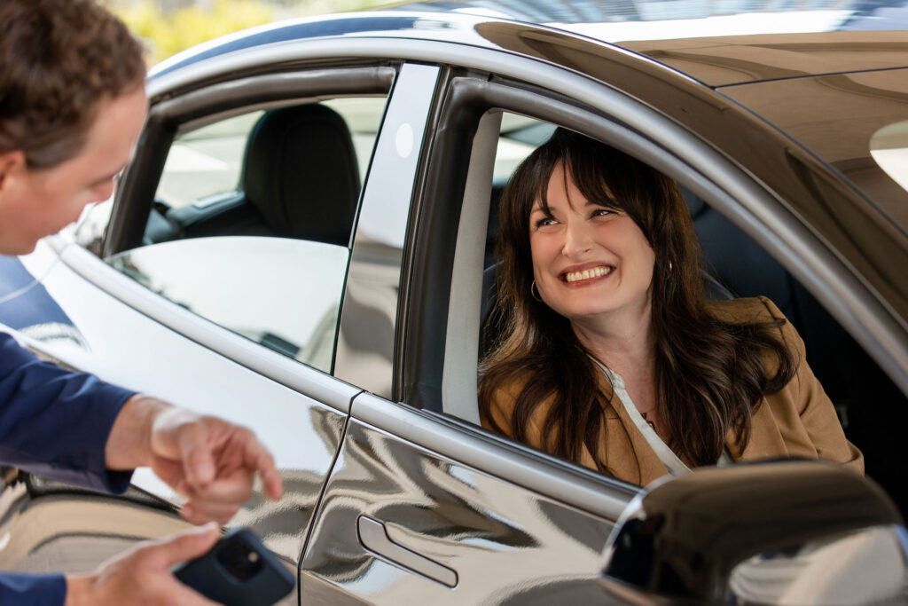 A woman sitting in the driver's seat of a car smiles through the open window at a man standing outside, who is holding a cell phone.