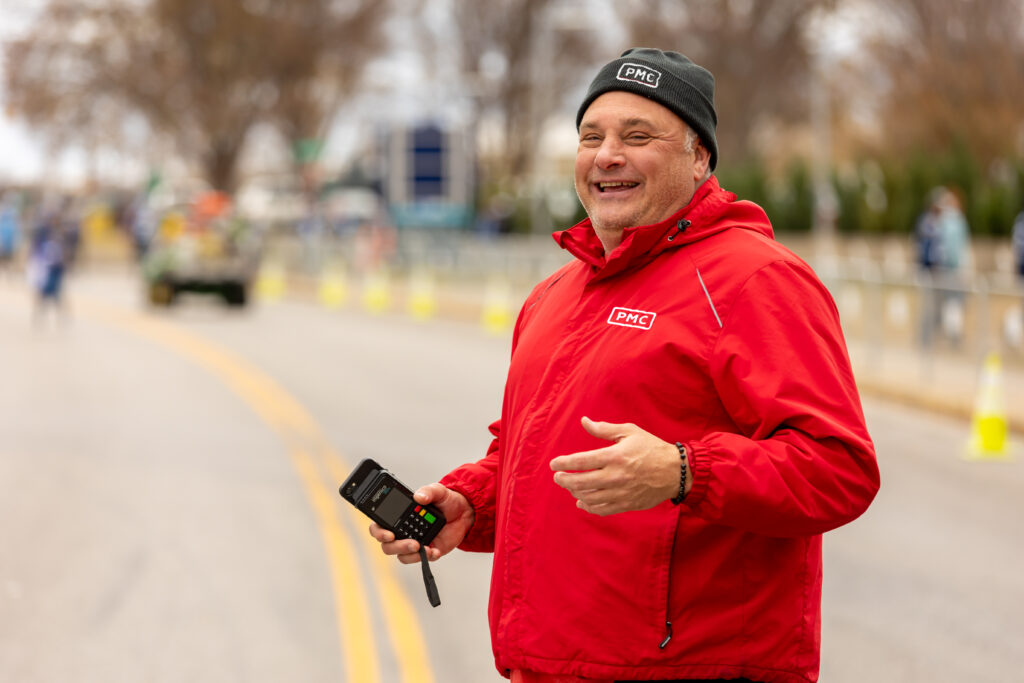 A man standing in the road in a red PMC jacket and black PMC beanie is smiling at the camera while holding a ticket scanner.