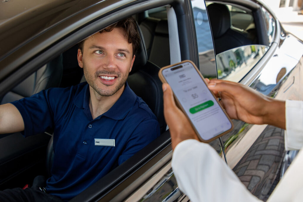 A valet driver sitting in a car with a navy PMC polo smiles at another person who is holding up a smartphone displaying a digital tipping app on the screen.