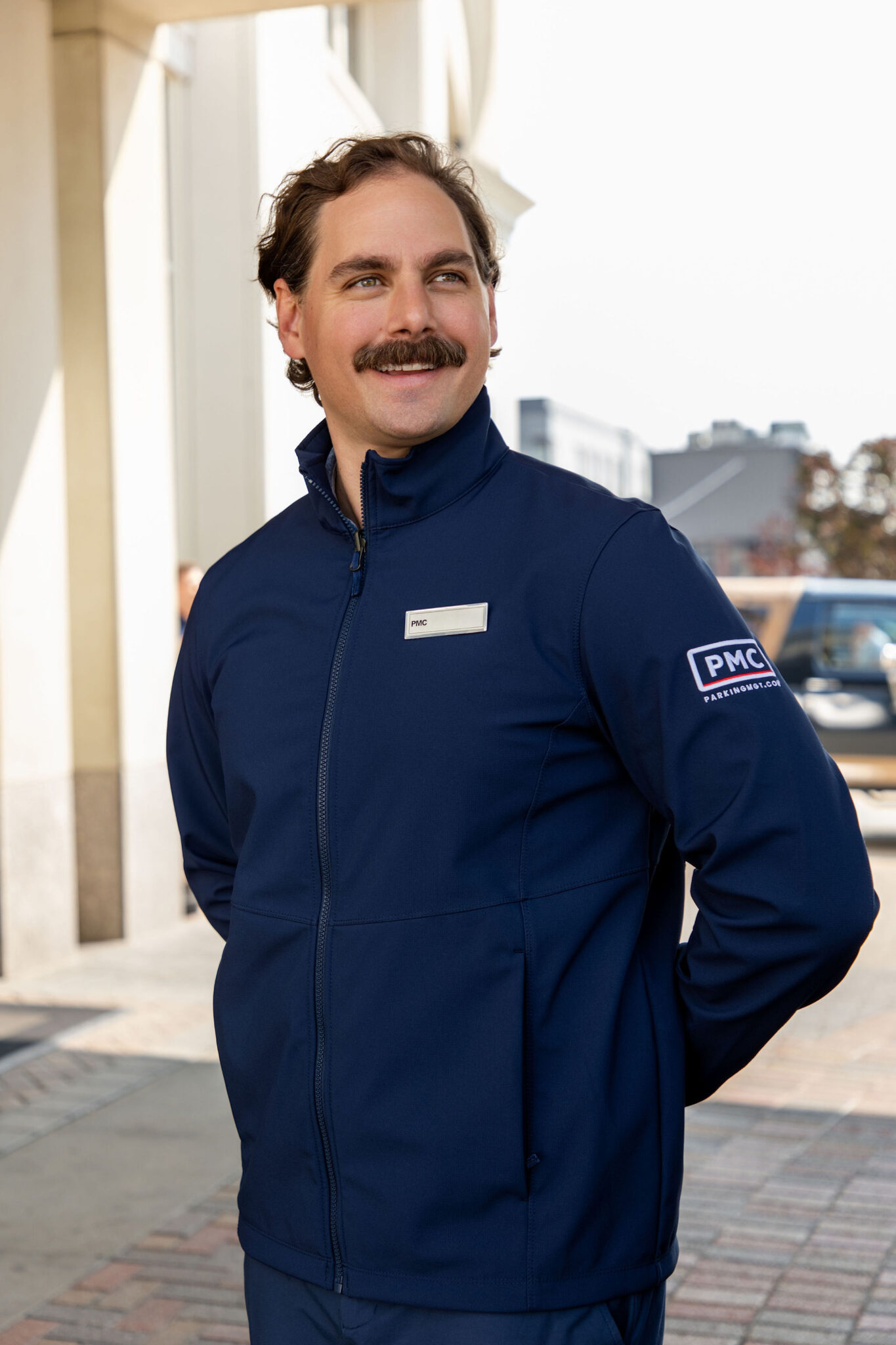 A man with brown hair and a mustache wears a navy blue jacket with a "PMC" patch on the sleeve and a name tag, standing outside on a sidewalk, smiling and looking to the side.