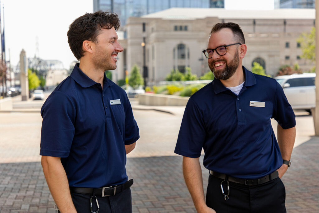 Two men wearing navy blue PMC uniforms and name badges stand outdoors on a city sidewalk, smiling and talking to each other. Both have short hair, and one wears glasses. Buildings and trees are visible in the background.