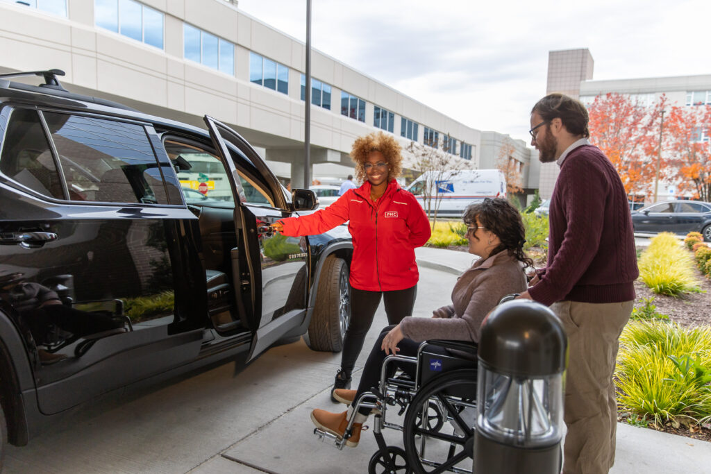 A woman in a red PMC jacket holds open a car door, smiling at a woman in a wheelchair and a man standing beside her outside a modern building on a cloudy day.