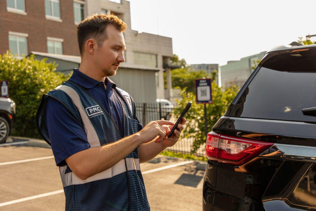 A parking attendant wearing a blue PMC vest uses a smartphone while standing next to a black SUV in a parking lot on a sunny day. Buildings and greenery are visible in the background.