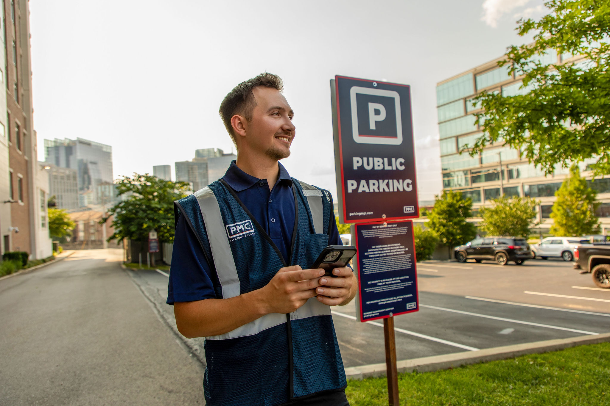 A smiling parking attendant in a blue PMC vest stands near a Parking Management Company sign, holding a phone, with an urban street and parked cars in the background on a sunny day.