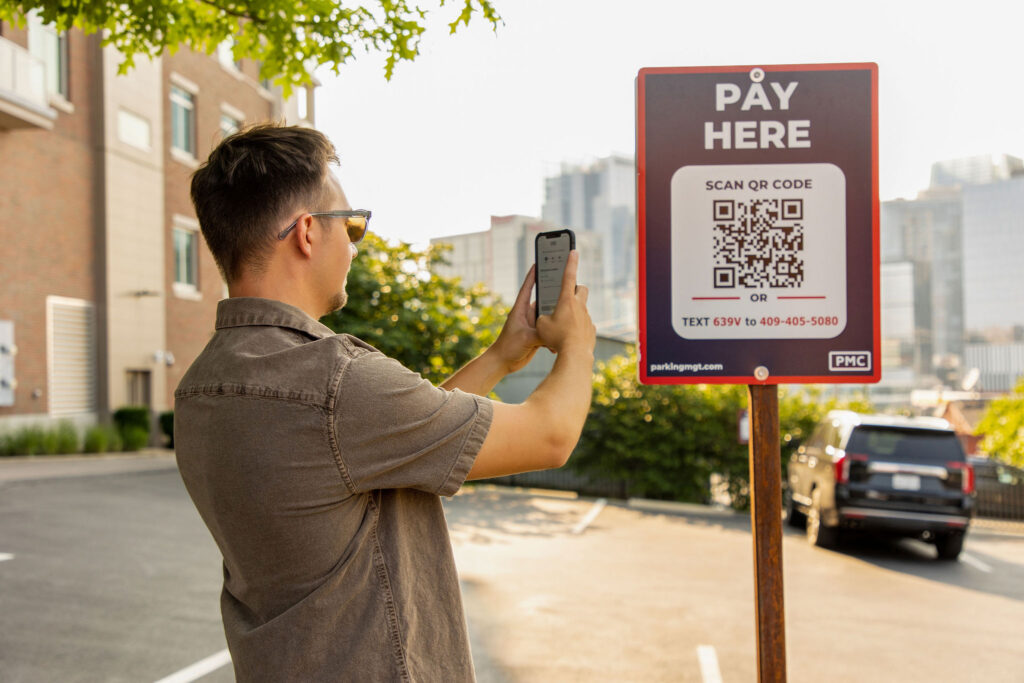 A man stands outdoors, holding his phone up to scan a QR code on a Parking Management Company parking sign. The city skyline is blurred in the background.