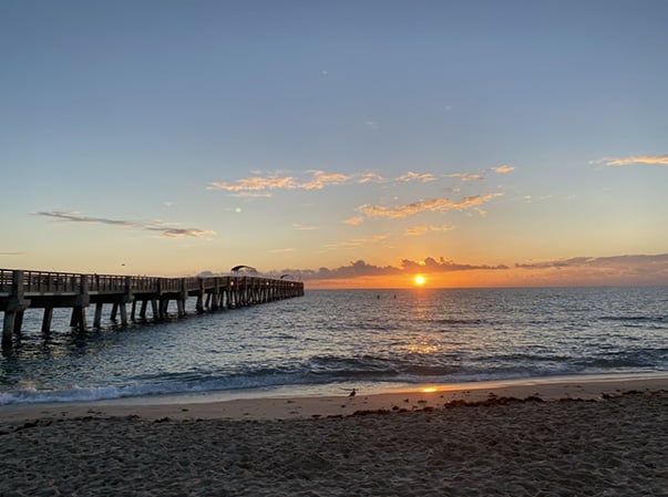 A long pier extends over calm ocean water at sunrise, with the sun low on the horizon, orange and pink clouds in the sky, and gentle waves washing onto the sandy beach in the foreground.
