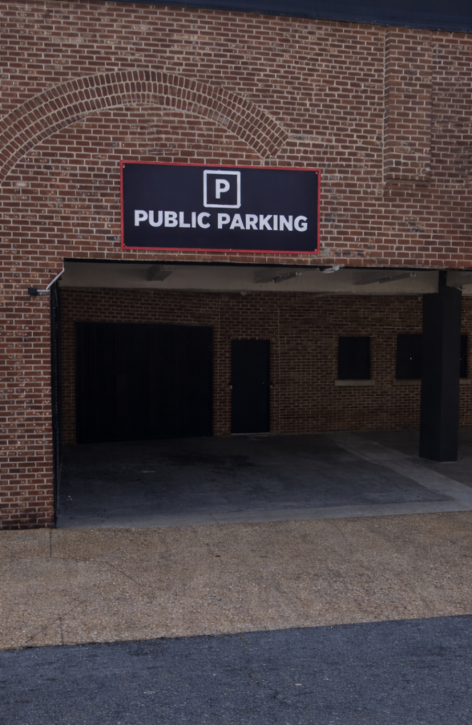 A brick building with an entrance to a covered parking area, marked by a black and white "Public Parking" sign above the entrance. The ground outside is paved with gravel and asphalt.