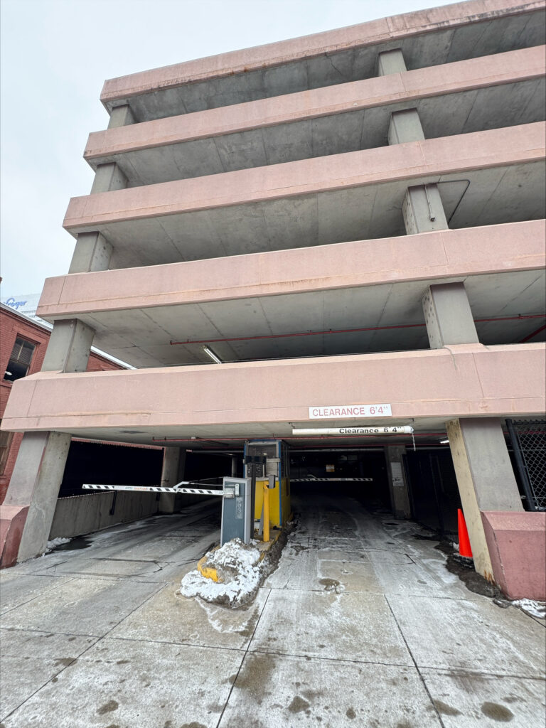 A multi-level concrete parking garage with an entrance gate, clearance sign reading "8' 4"," and some snow on the ground. A black gate and orange traffic cone are visible near the entrance.