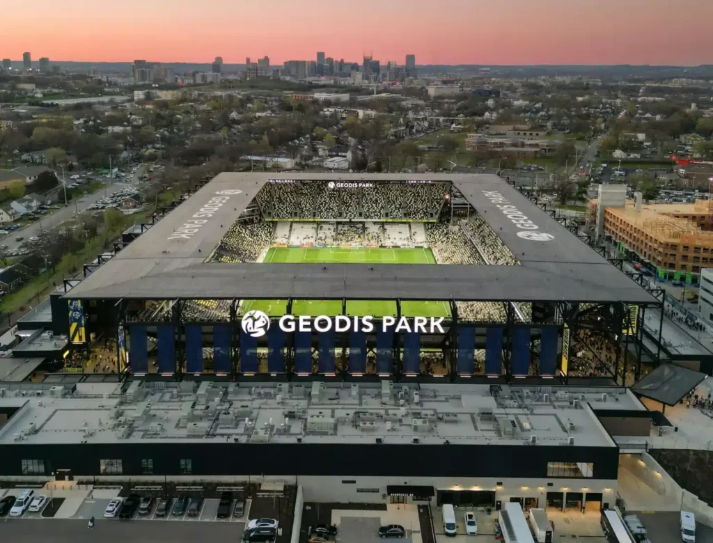 Aerial view of Geodis Park stadium at sunset, with the field illuminated and the city skyline in the background. Cars are parked around the stadium and trees and buildings surround the area.